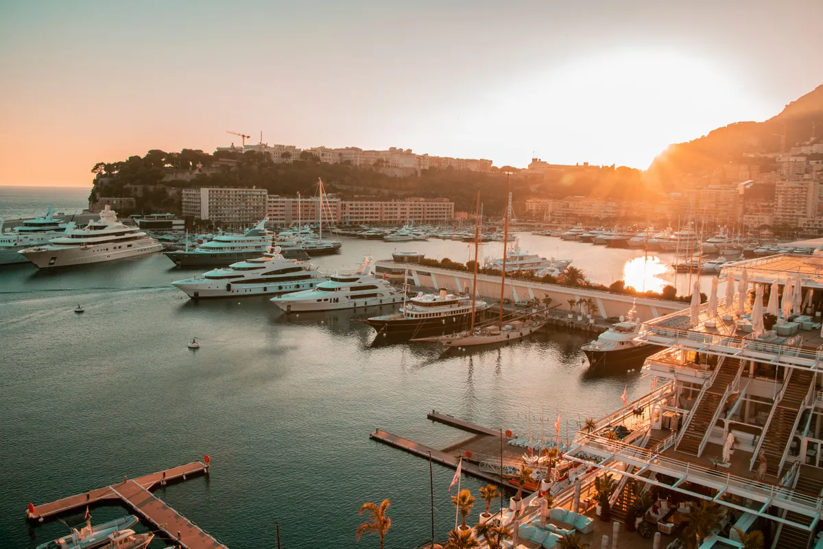 Yachts docked in a marina at sunset, with waterfront buildings and hills in the background.